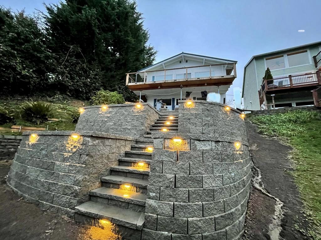 Curved block retaining wall with built-in lit staircase at dusk, built by 1st Choice Landscaping